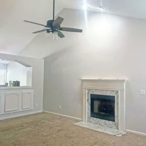 A low-angle view of a living room with vaulted ceilings and light gray walls. A fireplace with a white mantel and a gray and white marble surround is on the right side of the room. A dark brown ceiling fan with four wooden blades and a glass light fixture hangs from the ceiling. A half-wall with a built-in cabinet separates the living room from another space on the left. The floor is covered with a light brown, speckled carpet.
