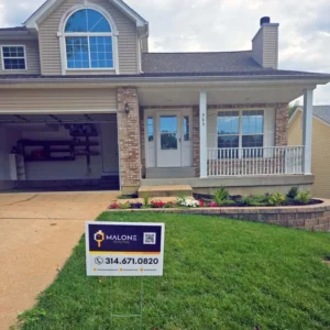 A two-story suburban house with a two-car garage. The house has a small porch with white columns and railings, and a flower bed in the front. A sign for "MALONE PAINTING" with a phone number and QR code is on the lawn. The sky is overcast.