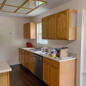 A galley kitchen with light wood cabinets and a stainless steel dishwasher. The floor is covered in dark wood. The countertop is a light, speckled color with a double sink and a faucet. Boxes and other items are cluttered on the countertop. A person with a ponytail and a white shirt is partially visible on the far left.