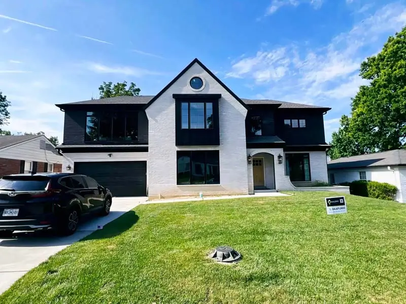 A two-story, modern-style house with a white brick facade and black siding. The house has a two-car garage, and a black SUV is parked in the driveway. A sign with the name "Malone" is visible on the green lawn. The sky is blue with some clouds.