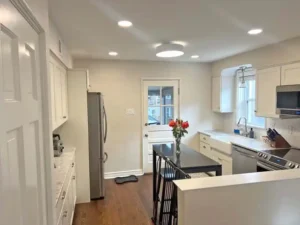 A white kitchen with dark wood floors and recessed lighting. A stainless steel refrigerator is on the left, next to a counter with a marble-like surface. A black island with three black stools is in the center of the room, with a vase of red flowers on top. A white farmhouse sink is in front of a window on the right, and a back door with a glass panel is visible in the background.