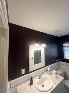 A modern bathroom with dark, possibly black or dark purple, walls. A white sink basin is set into a countertop with a gray and white marble pattern, and there is a matching tile backsplash. A white-framed medicine cabinet with a light fixture above it is mounted on the wall over the sink. A toilet is visible on the right.