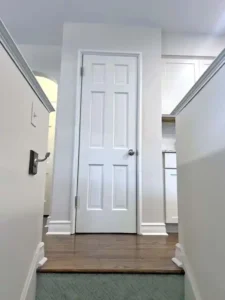 An indoor view from the bottom of a set of stairs. The stairs have a light-colored geometric patterned runner. At the top of the stairs, there is a landing with dark wood floors and a white six-panel door in the center. The walls on either side of the stairs are white and have white railings and trim. The hallway to the left of the door and a kitchen with white cabinets to the right are also visible.