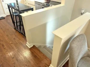 An elevated view of a split-level home's interior, showing a transition between a kitchen and a living area. A half-wall with a white ledge and baseboard molding separates a dark wood floor from carpeted stairs. A gray chair is visible in the foreground on the right, and a black kitchen island with a dark countertop is on the far left.