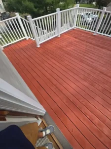 A wooden outdoor deck with a newly painted reddish-brown color, and white railings. A person's feet and legs are visible on the steps leading up to the deck.