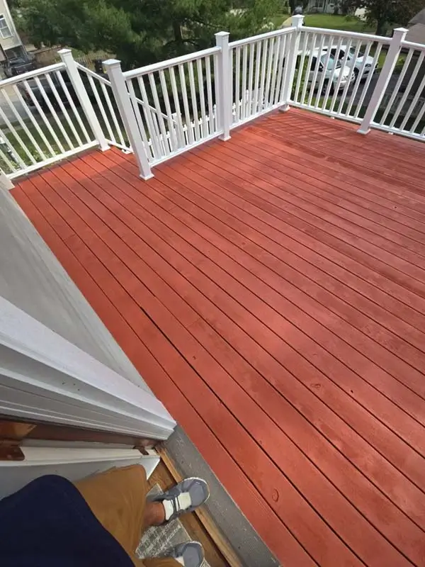 A wooden outdoor deck with a newly painted reddish-brown color, and white railings. A person's feet and legs are visible on the steps leading up to the deck.