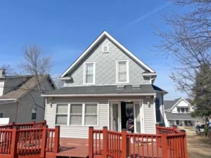A gray two-story house with a large red wooden deck in the foreground. The upper half of the house has scalloped siding and an A-frame roof, while the lower half has horizontal siding. There are several windows with white trim and a glass door leading to the deck. A person in a blue hoodie is standing near the doorway. The sky is bright blue with a white contrail.