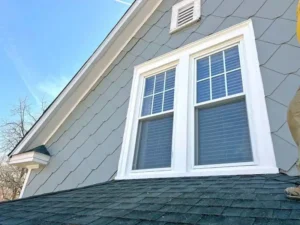 A close-up of the upper part of a house with light gray scalloped siding. There is a large white-framed double window with a grill on the top panes and blinds on the bottom. The dark shingled roof is visible below the window, and a white vent is near the peak of the house. A person's leg in khaki pants and a sneaker is visible on the right side of the image, standing on the roof.