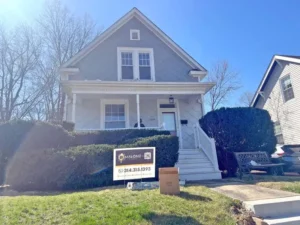 A three-story house with a blue exterior and white siding on the upper part. The house has a white porch and white railings on the stairs. A sign for "MALONE" with a phone number is on the lawn, and a cardboard box is near the sign. The sky is bright and sunny.