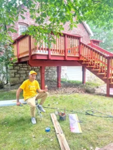 A red brick house with a wooden deck and a set of stairs leading to a backyard. A man in a yellow t-shirt and a yellow baseball cap is sitting on a metal piece on the ground, holding an ice cream cone. Various tools and pieces of wood are on the lawn. The sky is bright and sunny.