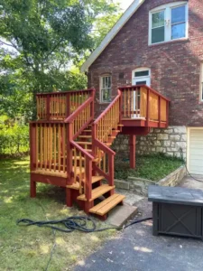 A multi-level, red wooden deck with stairs leading down to a paved area. The deck is attached to a two-story red brick house with a stone foundation. The top of the stairs and deck floors are a light, golden-brown wood color. A dark gray fire pit is visible on the paved ground in the foreground.