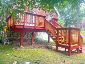 A raised red wooden deck attached to a red brick house with a stone foundation. The deck has a set of stairs leading down to a grassy yard. The railings are painted a solid red, while the top of the stairs and deck floor have a golden-brown finish. The yard is partially covered in scattered twigs and branches.