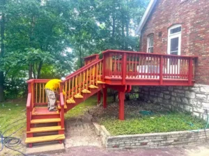 A side view of a red brick house with a raised wooden deck and stairs. The deck and railings are painted a deep red, while the top of the stairs are a golden yellow color. A person in a bright yellow shirt is working on the stairs near the bottom. The house's foundation is made of large, stacked stones, and there is a stone retaining wall in the foreground. Green trees and other foliage are visible in the background.