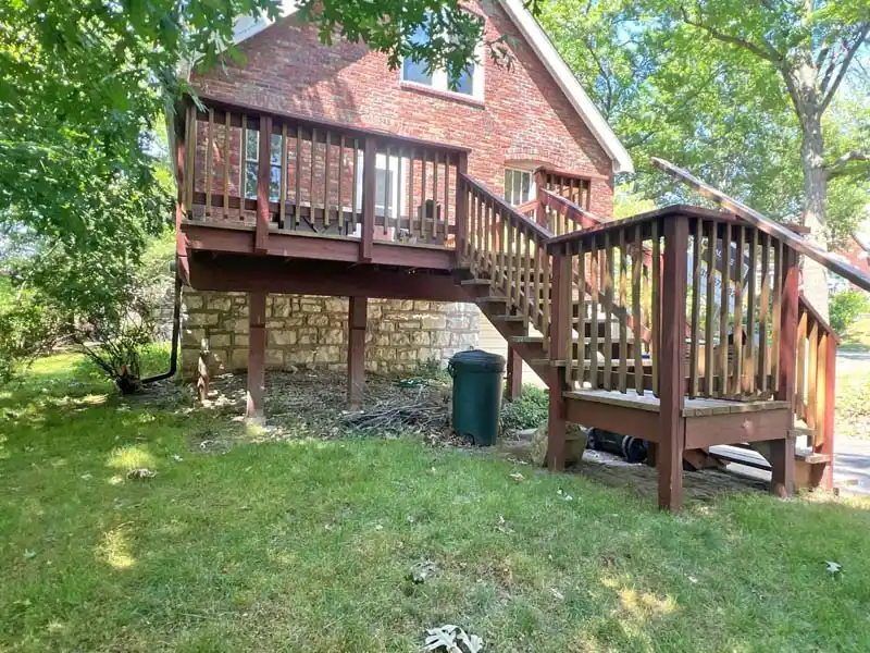 A back view of a red brick house with a raised wooden deck. The deck has a set of stairs leading down to a grassy yard. The house's foundation is made of large, stacked stones. A green trash can sits on the ground beneath the deck.