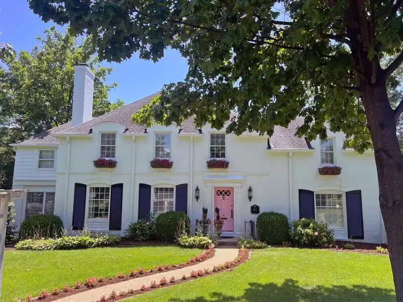 A white, two-story house with black shutters and a vibrant pink front door. The house has a manicured lawn with a curved walkway leading to the entrance, flanked by flower beds and lush green bushes. Several windows on the second floor have red flower boxes.