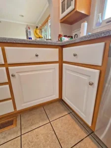 A close-up of kitchen cabinets with white doors and drawers, framed by light-colored wood. The countertops are a speckled gray, and the floor is made of large, light-brown tiles. A bunch of bananas and other items are visible on the counter.