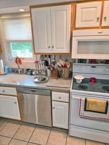 A small kitchen with white cabinets, a speckled gray countertop, and a tiled floor. There is a stainless steel dishwasher, a white stove, and a double sink under a window with a white blind. Kitchen utensils are hanging from a rail over the sink.