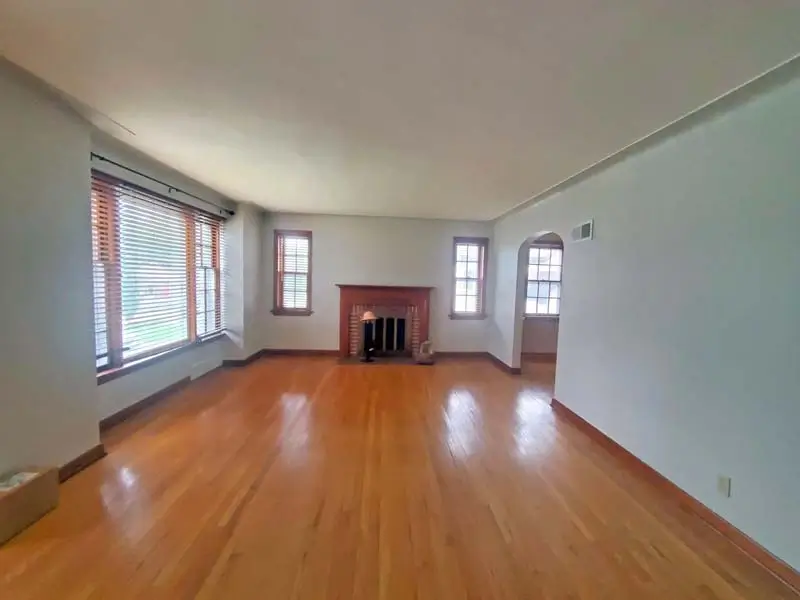 An empty living room with light gray walls and polished hardwood floors. The room features a fireplace with a dark wood mantle, and multiple windows with brown frames and wooden blinds. An arched doorway is visible on the right.