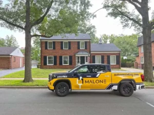 A bright yellow and navy blue pickup truck with a "MALONE PAINTING" logo is parked on the street in front of a two-story red brick house. The house has a black roof and black shutters on the windows. There are trees on both sides of the street and a small detached garage is visible on the left.