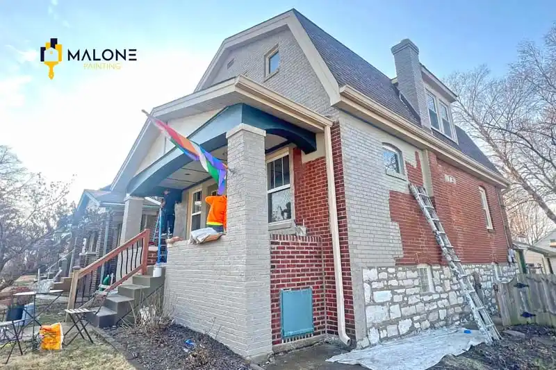 A two-story brick house is in the process of being painted from red to a light gray. The side of the house shows the unpainted red brick, while the front is a mix of painted gray and a light stone facade. Two workers are on a ladder and a porch, preparing to paint.