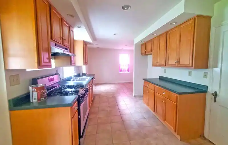 A view of an empty kitchen with light brown wood cabinets and green countertops. The room has light-colored tile floors and a stainless steel stove on the left side. The space appears to be in the middle of a renovation, as an open can of paint sits on the counter.