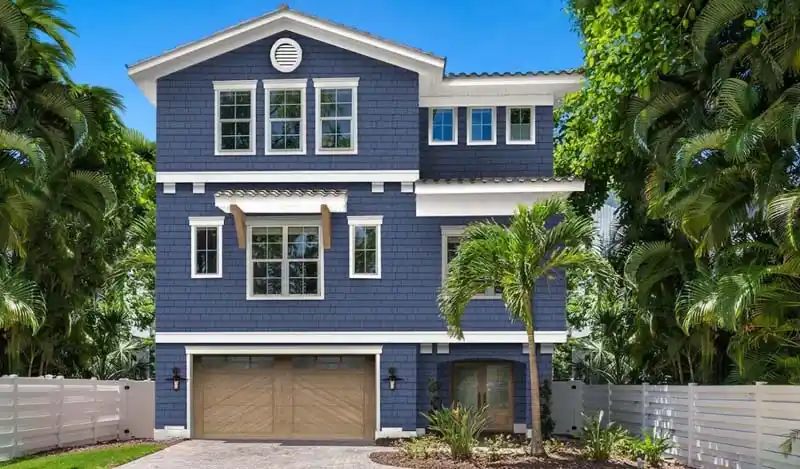 A large, two-story house with navy blue shingle siding and white trim. The house has a two-car garage with wood-paneled doors, a white fence in the foreground, and is surrounded by lush tropical trees and bushes. A small palm tree is in the front yard.