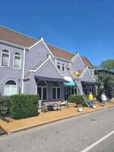 A long, light purple commercial building with multiple gables and a brown roof. A person is on a ladder on the roof, and two other people are on the sidewalk below. The building has several awnings and outdoor seating on a patio. The sky is clear blue.