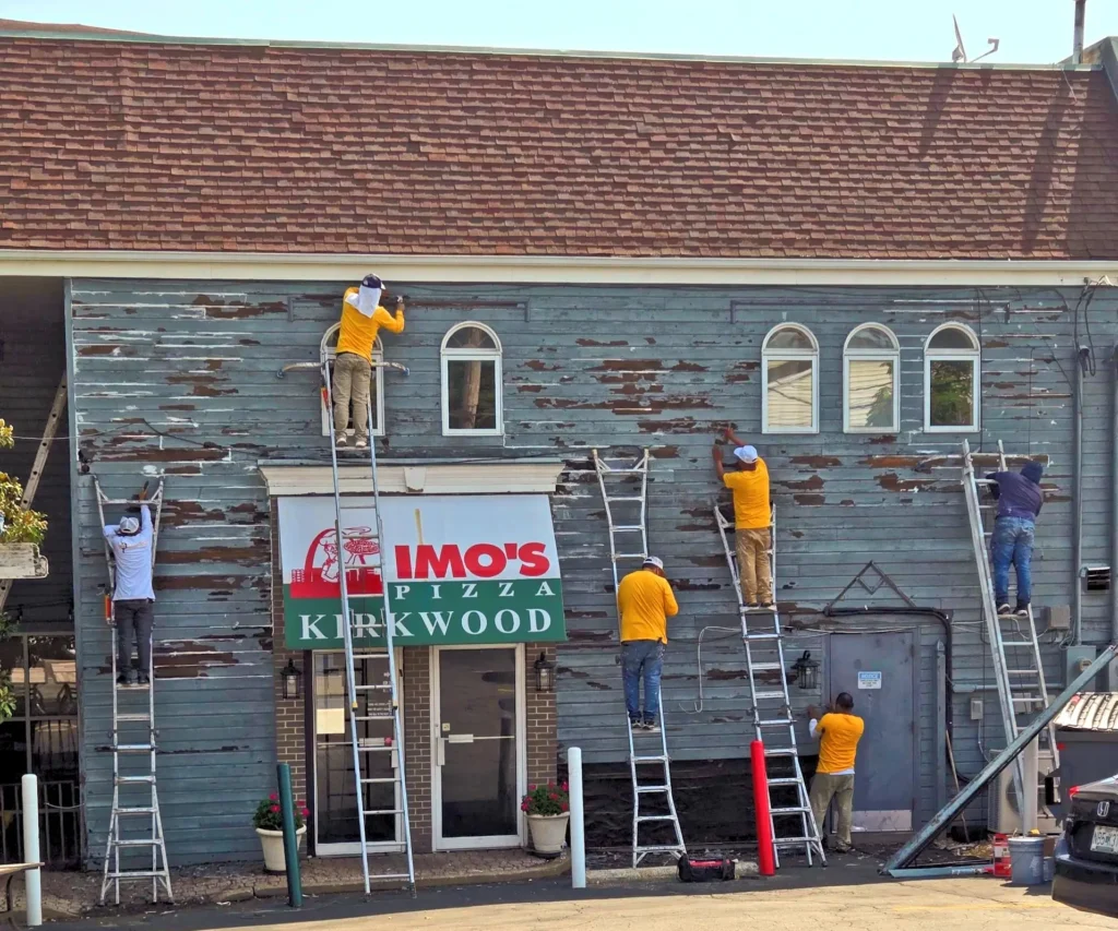 An outdoor shot of the front of a two-story building. The building has a reddish-brown shingled roof and light blue siding that is peeling in many places, revealing the bare wood underneath. The sign for "Imo's Pizza Kirkwood" is visible on the first story, above a glass door entrance. Five people are on ladders, actively working on the exterior of the building. Most of them are wearing yellow shirts and white hard hats. The windows on the second story are arched at the top