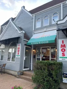 A light gray two-story building with a sign for "Imos Pizza" on the front. A green awning with "KIRKWOOD" written on it is over the main entrance. The building has multiple windows and a small hedge in front of it.