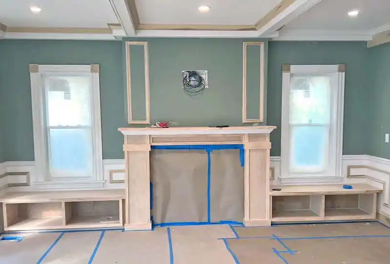 A living room undergoing renovation, with a partially built wooden fireplace mantel and built-in benches under two windows. The walls are a light green color with white and unpainted wood trim. The floor is covered in brown paper and blue painter's tape, and wires hang from the wall above the fireplace.