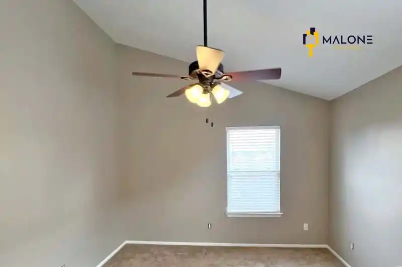 A high-angled view of a room with a vaulted ceiling and a ceiling fan. The walls are a light beige color, a window with blinds is on the far wall, and the floor is covered in a brown carpet. A small Malone Painting logo is in the top right corner.