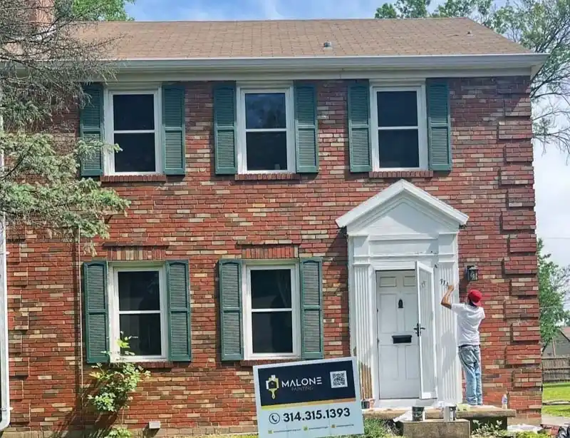 A worker paints the white trim around the front door of a two-story brick house with green shutters. A Malone Painting sign with a phone number and QR code is visible in the foreground on the left.