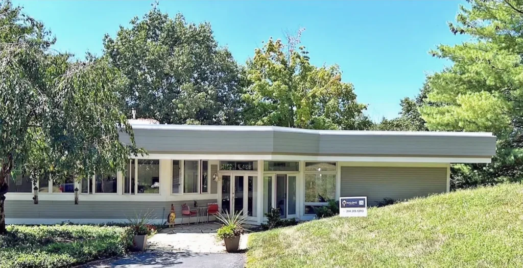 A single-story, mid-century modern home with sage green siding and a distinctive flat roof. The house features large walls of windows and is built into the side of a grassy hill, with tall green trees in the background
