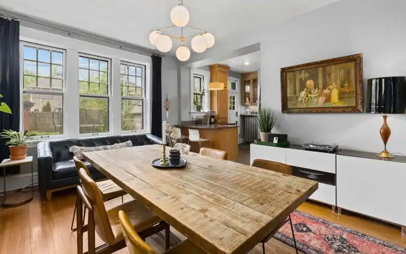 A bright dining room with light gray walls, a hardwood floor, and a large wooden dining table with leather chairs. A black leather couch is placed under a large window with black curtains. A modern white cabinet with a record player sits against the wall, and an antique painting hangs above it. In the background, an open doorway leads into a kitchen.
