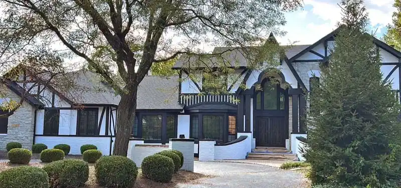 A large, two-story Tudor-style house with a light gray stone facade and white stucco with dark brown timber framing. The house has a black front door, a prominent second-story balcony, and a driveway that curves past a series of neatly trimmed shrubs in the front yard.