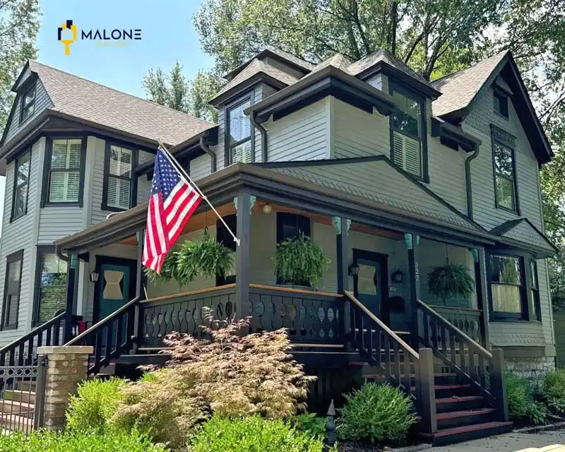 A modern Victorian-style house with a light gray siding and a dark roof. The house has a two-story porch with intricate black railings and a green front door. An American flag hangs from the porch, and several hanging planters are visible. A small Malone Painting logo is in the top right corner.