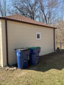 A low, single-story house with light yellow and brown siding and a brown roof. Two large blue trash cans with black and green lids are in the yard next to the house. The sky is bright blue with no clouds.
