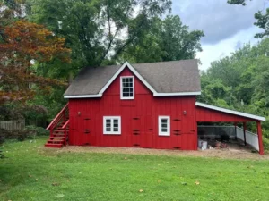 A small, two-story red barn with a brown shingle roof and white trim. A set of red wooden stairs leads to the upper level on the left. The building has a covered lean-to on the right side where some paint cans are visible. The barn is in a grassy yard surrounded by green trees.