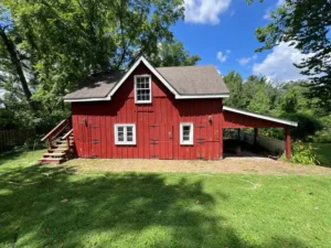 A small, red barn-like building with a brown roof and white trim is shown in a grassy yard. The building has a covered lean-to on the right side and a set of wooden stairs leading to an upper level on the left. The sky is bright blue with white clouds, and the building is surrounded by green trees.