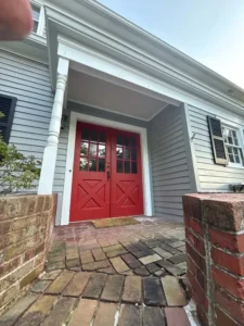 A low-angle view of a house with light gray siding and a bright red double front door with window panes. The door is under a white portico supported by two large white columns. The walkway leading to the door is made of mismatched red bricks and is flanked by two low brick walls. Black shutters are visible on the windows to the side of the door.