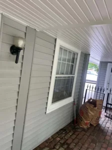 A low-angle view of a house exterior with light gray siding and a white window frame with panes. The ceiling of the porch is white and paneled, and the floor is made of red brick. A black light fixture is on the wall, and two large brown paper bags are leaning against a black metal fence on the right.