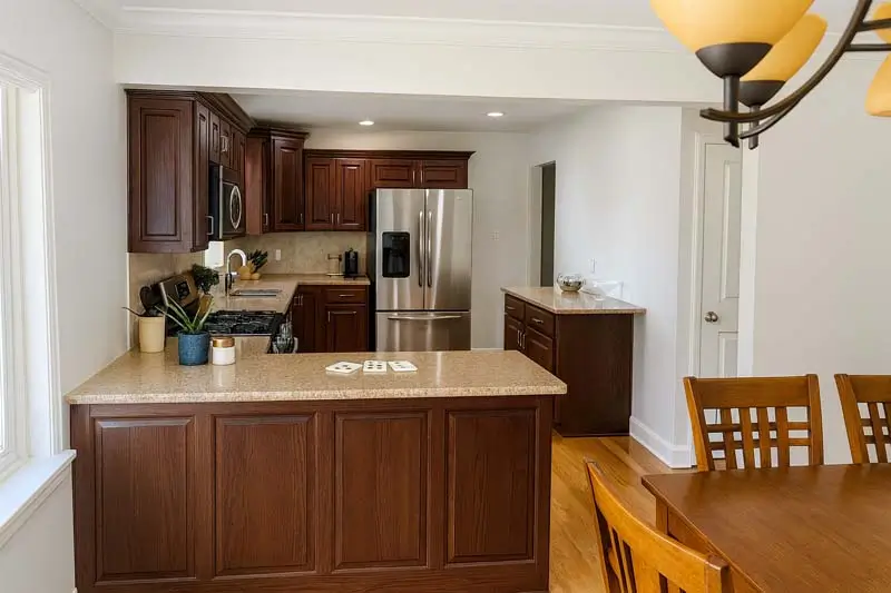 A modern kitchen with dark brown wooden cabinets, light-colored countertops, and hardwood floors. The kitchen has stainless steel appliances, including a double-door refrigerator. An island with two teal pots is in the foreground. A dining room table with chairs is visible on the right side of the image.
