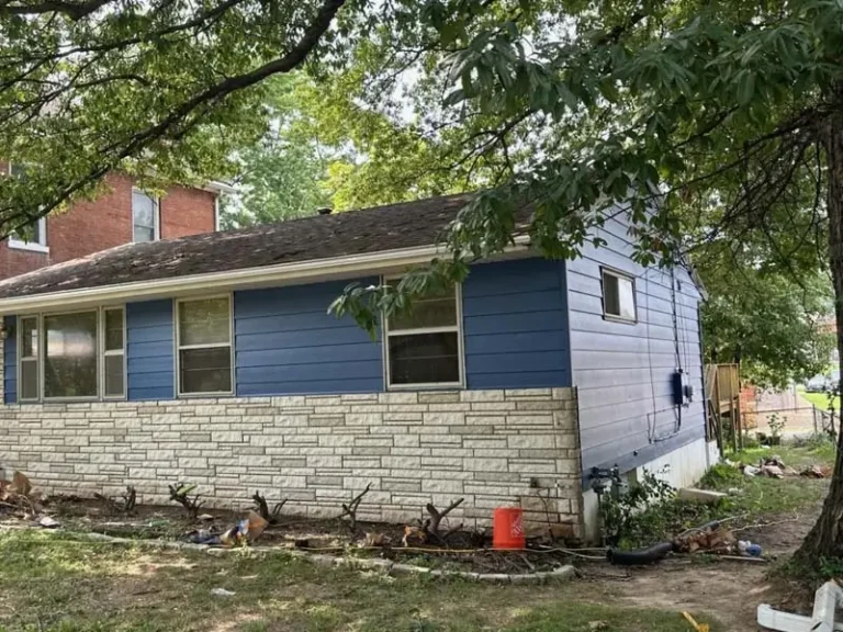An outdoor shot of a single-story house with light blue siding on the top portion and a light-colored, stone facade on the bottom. The front yard is in a state of renovation, with exposed dirt and a few small shrubs in a planter bed. A large tree with a thick trunk is visible on the right, and its branches hang over the house. A red brick house is partially visible in the background to the left. A red bucket is sitting near the front of the house