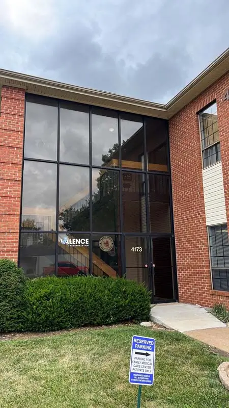 A red brick office building with a large, two-story glass facade and a black front door. The number "4173" is visible on the glass. A sign for "VALENCE" is on the inside of the window, and another sign for "RESERVED PARKING" is on a post in the foreground. Bushes are planted along the base of the building.