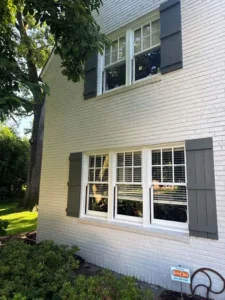 A horizontal, close-up view of the side of a two-story white brick house. There are two sets of white-framed windows with dark gray shutters. A large, leafy tree is on the left side of the frame. In the foreground, there are green bushes and a small white and red sign in the grass.