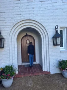 A straight-on view of the front door of a white brick house. The door is made of dark wood and has a curved top, and it is set back in an arched entryway with a red tile floor. A man in a black jacket and blue jeans is standing in front of the door. A black light fixture hangs on the wall to the right of the door. There are two potted plants with pink flowers on either side of the entrance.