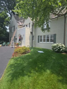 A horizontal view of a two-story light-colored brick house. The house has dark gray shutters and a white roof. There is a small patio with furniture and a ladder near the front door. The house is surrounded by a well-maintained green lawn with a large bush and red and white flowers.