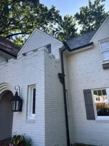 A vertical, low-angle shot of the corner of a two-story white brick house. The house has a complex roofline and a dark gray gutter system. A dark gray light fixture hangs in an arched entry on the first floor, and a dark gray shutter is on a window on the right. A large tree with green leaves is in the background.