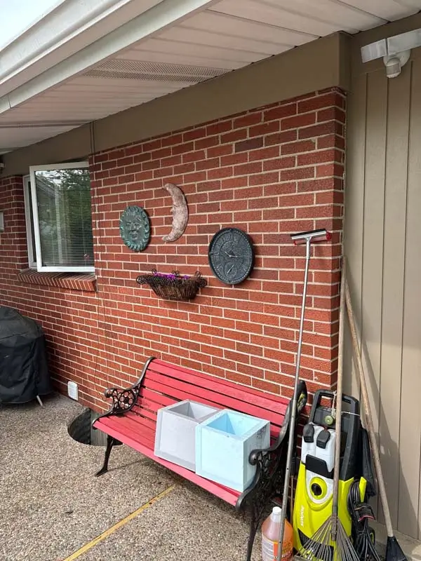 A small patio with a red brick wall. A red and black metal bench is pushed up against the wall, with two light-colored planters on it. A variety of yard tools, including a rake and a yellow pressure washer, are propped up against the wall on the right. Decorative items, including a sun and moon sculpture and a wall clock, are hanging on the brick.