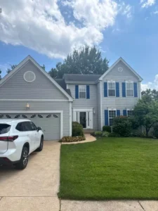 A two-story light gray house with a gray shingle roof and a two-car garage. The house has blue shutters on the windows and a white front door. A white SUV is parked in the driveway on the left. The lawn is green and well-maintained, and the sky is blue with scattered clouds.